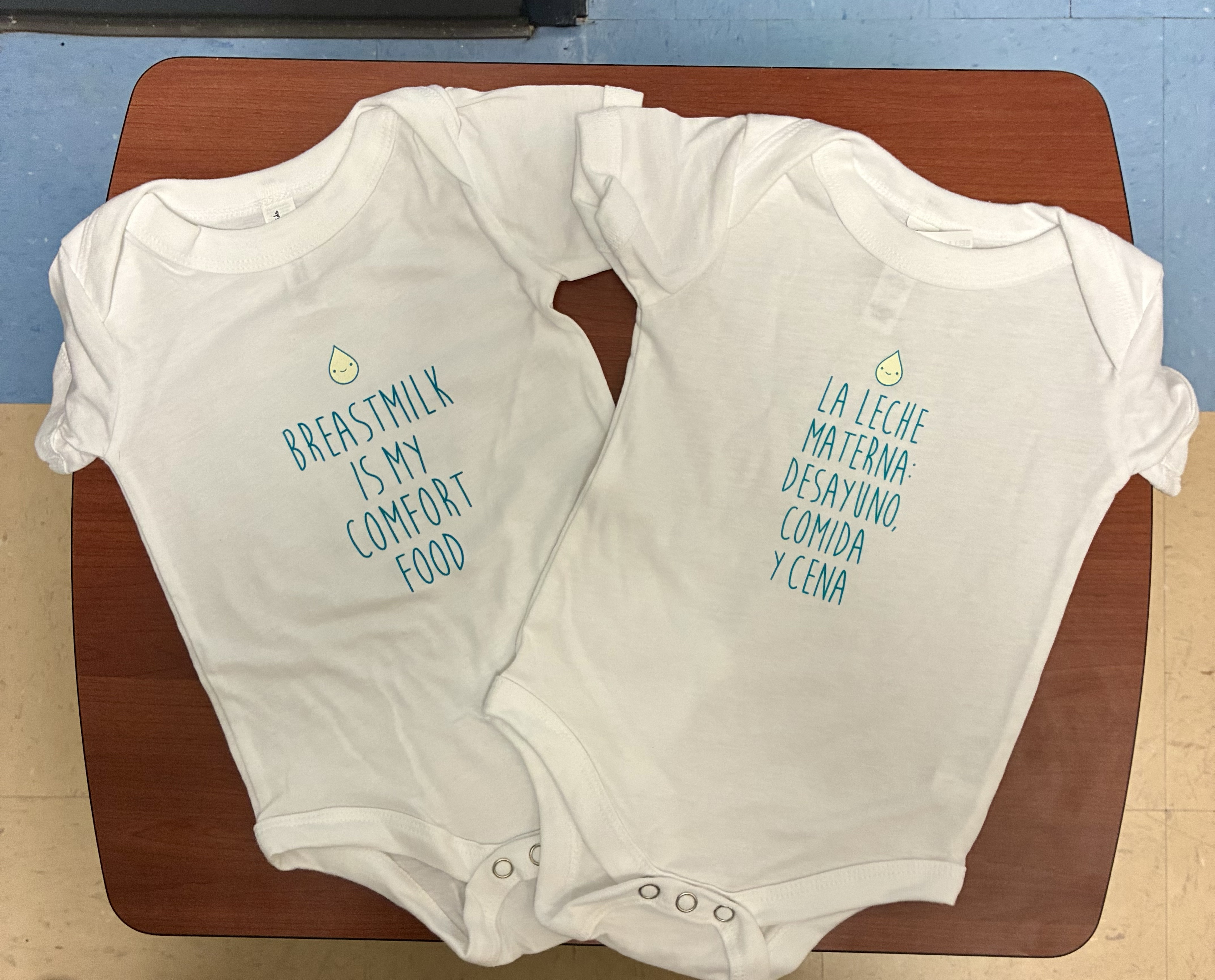 Two baby onsies sit side by side on a chair with English and Spanish imprint.